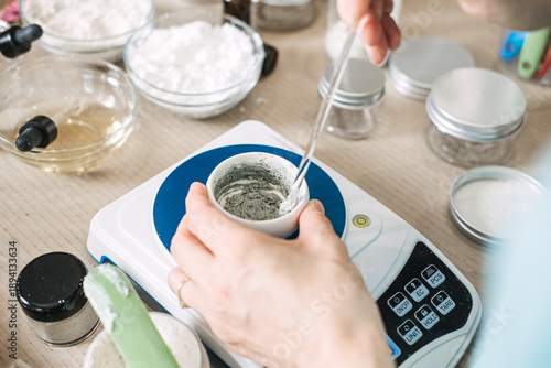 Hands stir gray cosmetic paste in small bowl on digital scale surrounded by laboratory glassware and jars. Small-batch manufacturing, slow beauty movement, artisan production.