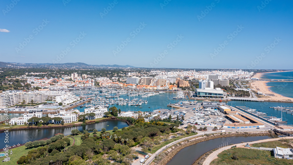Obraz premium A wide view shows Vilamoura marina with boats docked, people enjoying the area, and buildings lining the waterfront under a blue sky Aerial