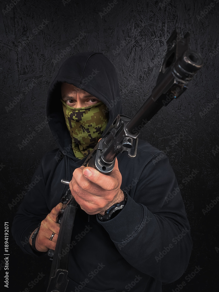 Obraz premium Armed man in black hoodie and camouflage mask holding a rifle against a dark background, representing security, tactical concept, or soldier.