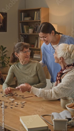 Vertical shot of young female nurse monitoring group of focused seniors assembling puzzle inside retirement home
