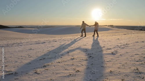 Young couple walks casting long shadows on snow at winter sunset. Romantic man and woman join hands during promenade on frozen valley. Happy tourists enjoy cold dusk in nature