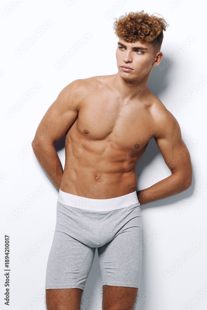 Obraz premium Model, man, young and athlete wearing gray boxer shorts with white elastic, posing shirtless with toned muscles and curly hair against white background in studio for fitness and health concept.