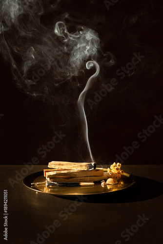 Bâtons de Palo Santo posés sur un plateau en bronze, un bâton allumé avec une fumée blanche qui s'élève vers le haut finissant en volute, fond brun foncé, ambiance méditative et relaxante