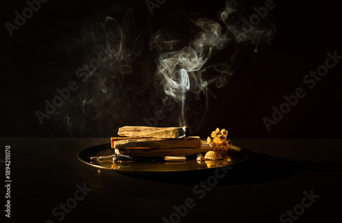 Bâtons de Palo Santo en fagot posés sur un plateau en bronze, un bâton sur le dessus brûlant et libérant une fumée blanche verticale, fond brun foncé, ambiance méditative et douce