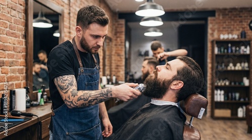 Professional male barber with tattoos and apron trimming beard of client in modern barbershop. Hairdresser using electric clippers. Men grooming concept. Stylish haircut salon