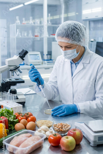 Female food scientist in white lab coat, gloves,mask and hair net using pipette to analyze food samples in modern lab. Professional researcher conducting quality control tests with fresh produce.