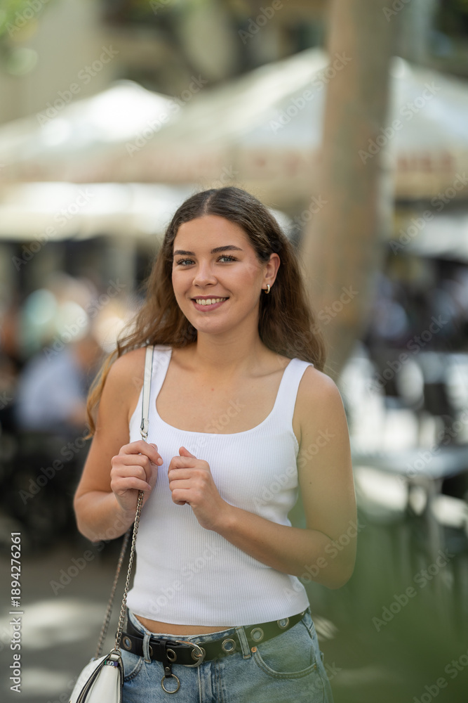 Obraz premium Young woman smiling and walks along sunlit street, walks around city, spends time in public urban space. Person and megalopolis..