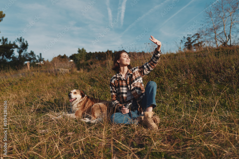 Fototapeta premium Woman in plaid shirt sits in a grassy field with a dog beside her, raising her arm toward the bright sky during a sunny outdoor moment in nature today.