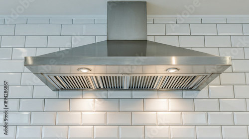 Modern stainless steel range hood hangs over white subway tile backsplash. Captures smoke and odors from cooking area. Kitchen appliance for ventilation. Essential home or restaurant item.