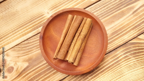 Cinnamon sticks are neatly arranged on a round, wooden plate placed on a wooden surface. The cinnamon sticks are a rich brown color, with a smooth texture and characteristic curled shape.