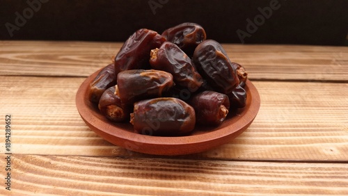 Dates are arranged on a round wooden plate resting on a wooden table.  dates have a glossy surface and rich, dark brown color, indicating ripeness. The wooden plate has a smooth finish 