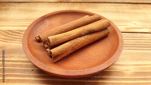Cinnamon sticks are neatly arranged on a round, wooden plate placed on a wooden surface. The cinnamon sticks are a rich brown color, with a smooth texture and characteristic curled shape.