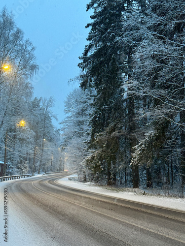 Snowy asphalt road winds through forest, lined with snow covered trees. Streetlights cast a warm glow on wintry scene. Tranquil winter evening. Vertical photography.