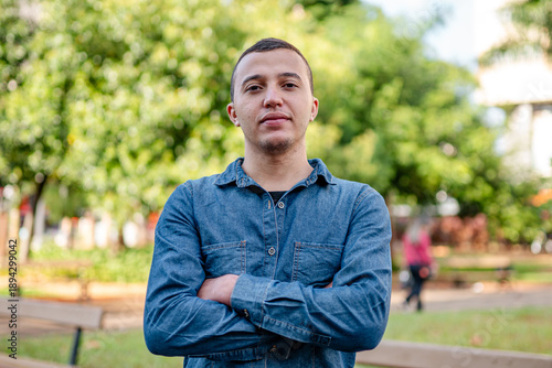Confident young man in blue denim shirt standing with arms crossed in a public park.