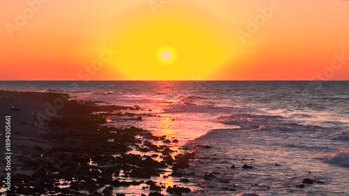 AERIAL, LENS FLARE: Peaceful evening seascape with setting sun above horizon, painting the sky orange and gold. Gentle waves wash over dark coastal rocks, creating shimmering reflections across sea.