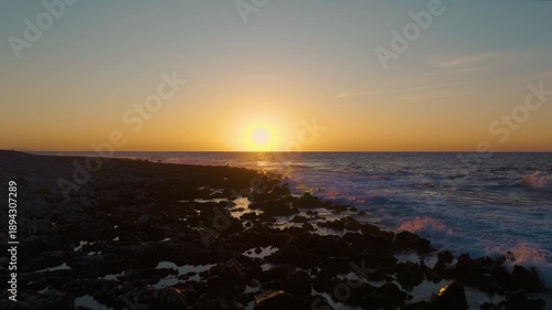 AERIAL, LENS FLARE: Rugged rocky shoreline and setting sun over the open sea. Warm golden light reflects in scattered puddles and gentle waves roll to the beach. Peaceful coastal landscape in Sicily