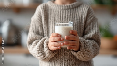 Kid holding glass of milk with both hands, bright kitchen background out of focus, photorealistic