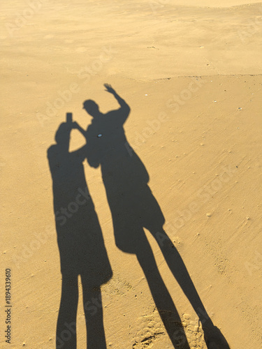 Silhouettes of two people on a smooth golden sandy beach waving and taking a picture of themselves on holiday having fun