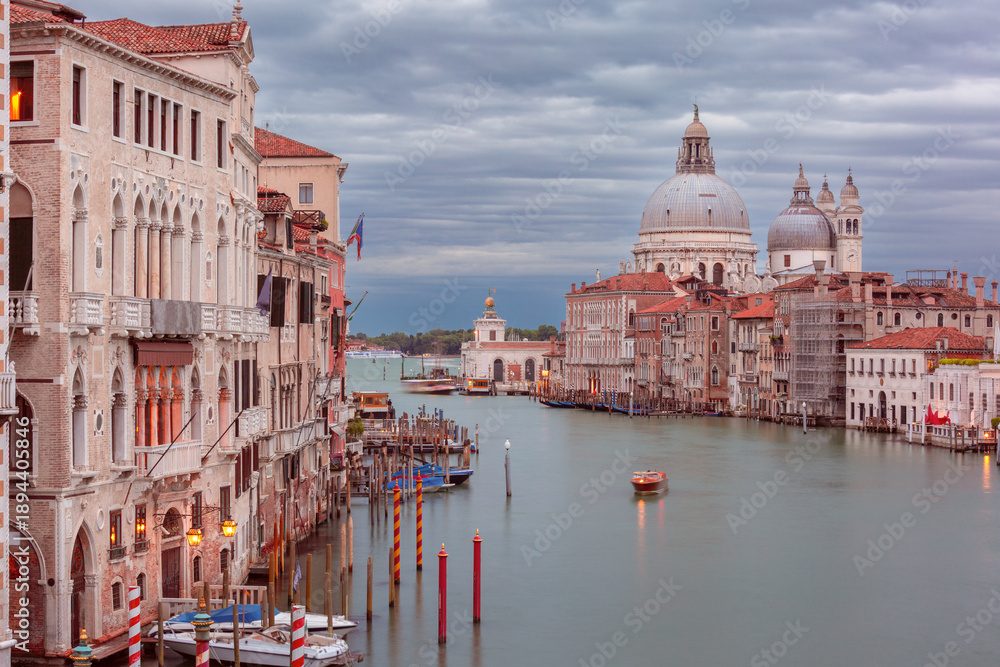 Naklejka premium A view of the Grand Canal in Venice, Italy with boats, historic buildings, and the Basilica di Santa Maria della Salute under a cloudy sky.