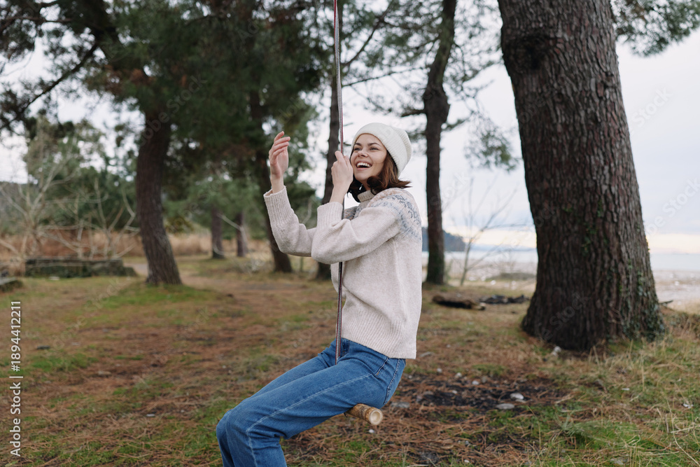 Fototapeta premium Young girl on a swing beside tall trees in a coastal forest, wearing a knit sweater and beanie, capturing a playful moment in nature.