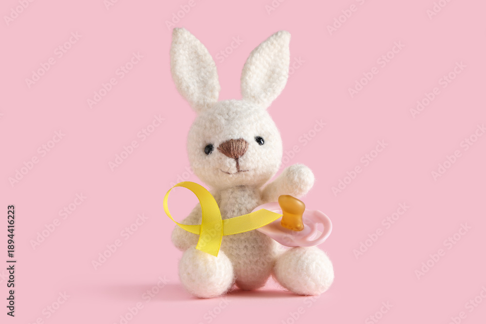 Obraz premium Toy bunny, pacifier and yellow ribbon on pink background. Childhood cancer awareness concept