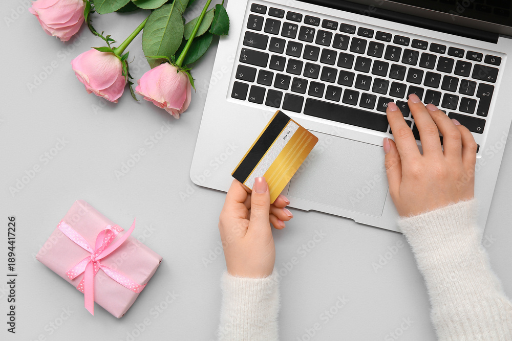 Fototapeta premium Female hands with credit card, modern laptop, gift and rose flowers for Valentine's Day on grey background, closeup