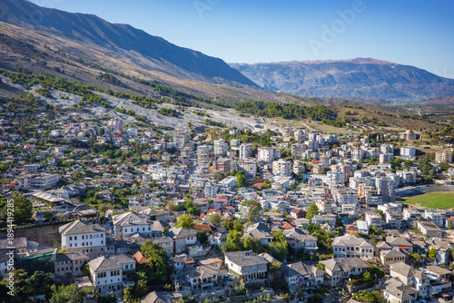 Wide panoramic view of a picturesque European town spread across a hillside and valley, showcasing historic stone-roof houses in the foreground and modern apartment blocks in the background