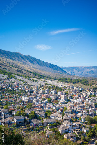 Wide aerial-style view of a vibrant city nestled at the base of rugged mountains, featuring dense residential neighborhoods, modern apartment buildings, and traditional rooftops surrounded by greenery
