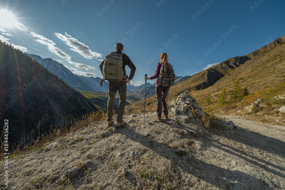 Obraz premium A male and female hiker stand on a high, rocky viewpoint under a bright sun and blue sky, looking out at a vast mountain range and a green valley during an autumn or summer trek.