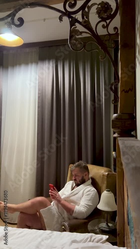 Vertical 4K video. 60 fps. Mature man with beard, white robe sits on chair in hotel bed room, watching social media news, message on phone. Concept of men day, call, wash, skin care, rest, relax, talk