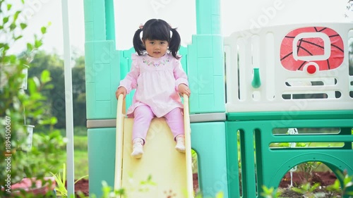 Happy toddler sliding down playground slide in bright sunlight, enjoying outdoor playtime with a joyful expression and innocence in a lush green environment