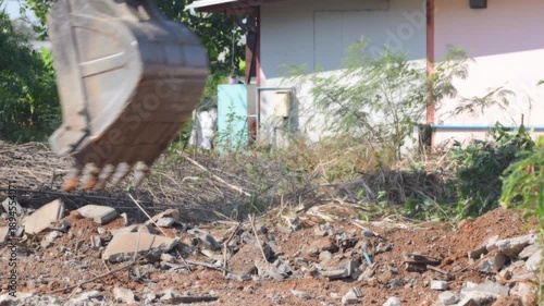 Excavator bucket digging soil, demolition rubble, construction debris cleanup at residential site, clearing broken concrete, brick, metal scrap, dusty earthmoving machinery after renovation