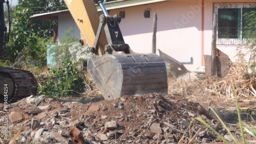 Excavator bucket demolition rubble construction debris earth house, heavy equipment clearing broken concrete and dusty soil beside small residential home during renovation site cleanup work