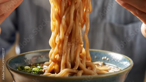 Hands use chopsticks to lift thick noodles from a ceramic bowl of broth garnished with green onions