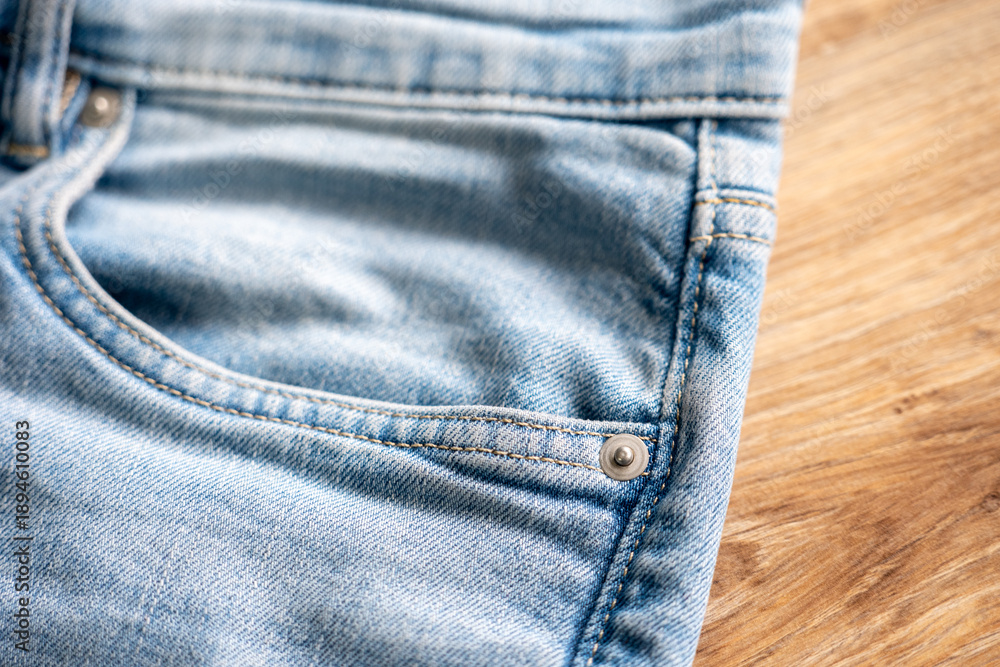 Fototapeta premium Close-up of blue denim jeans front pocket with metal rivet and stitching on wooden surface, suitable for fashion design, clothing detail, textile background, and casual wear concepts.
