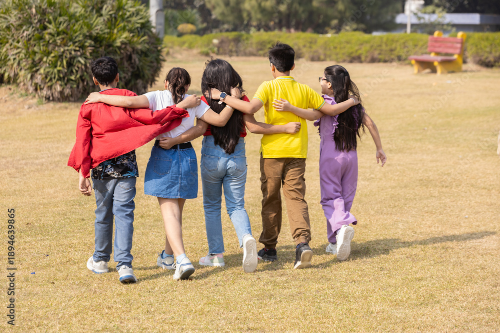 Obraz premium Group of indian children friends walking with arms around each other enjoying time together outdoor in summer park. Asian kids having fun. Childhood. Back view.