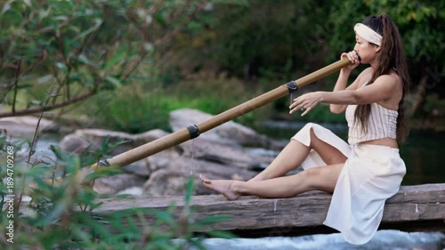 A serene moment by the water as a woman skillfully plays a bamboo Instrument didgeridoo, showcasing both grace and musicianship in a natural outdoor setting.