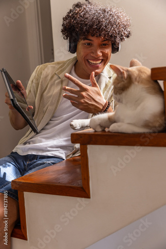Man sitting on staircase using tablet and headphones while cat basking in sunlight