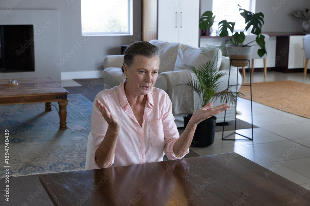 Fototapeta premium Senior woman gesturing with hands while talking at dining table in living room next to plants