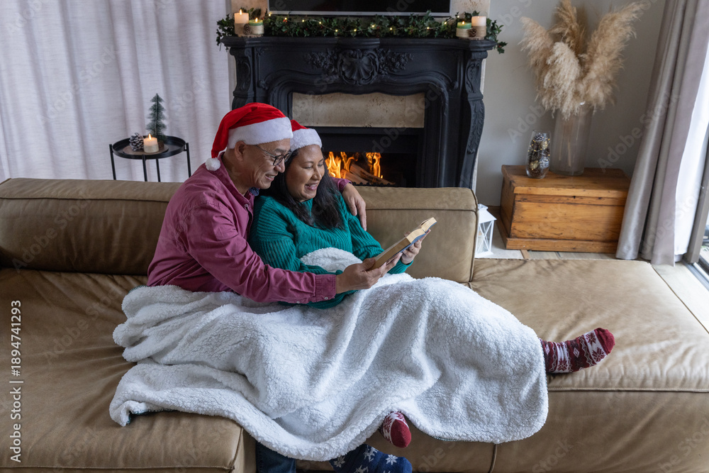 Fototapeta premium Senior couple sitting on leather couch reading book under white blanket by fireplace in living room