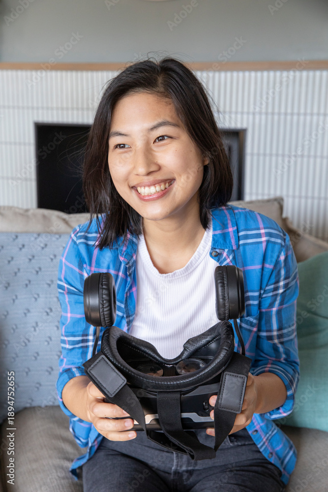 Fototapeta premium Asian woman sitting on grey couch in living room by fireplace holding virtual reality headset