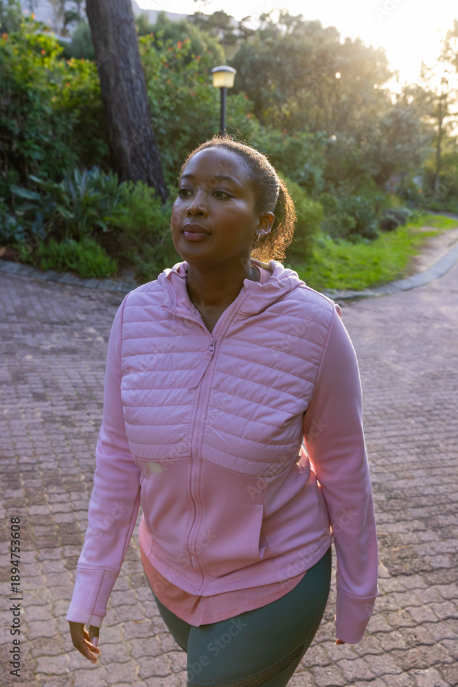 Fototapeta premium African american woman walking in activewear along brick path in park using wireless earbuds