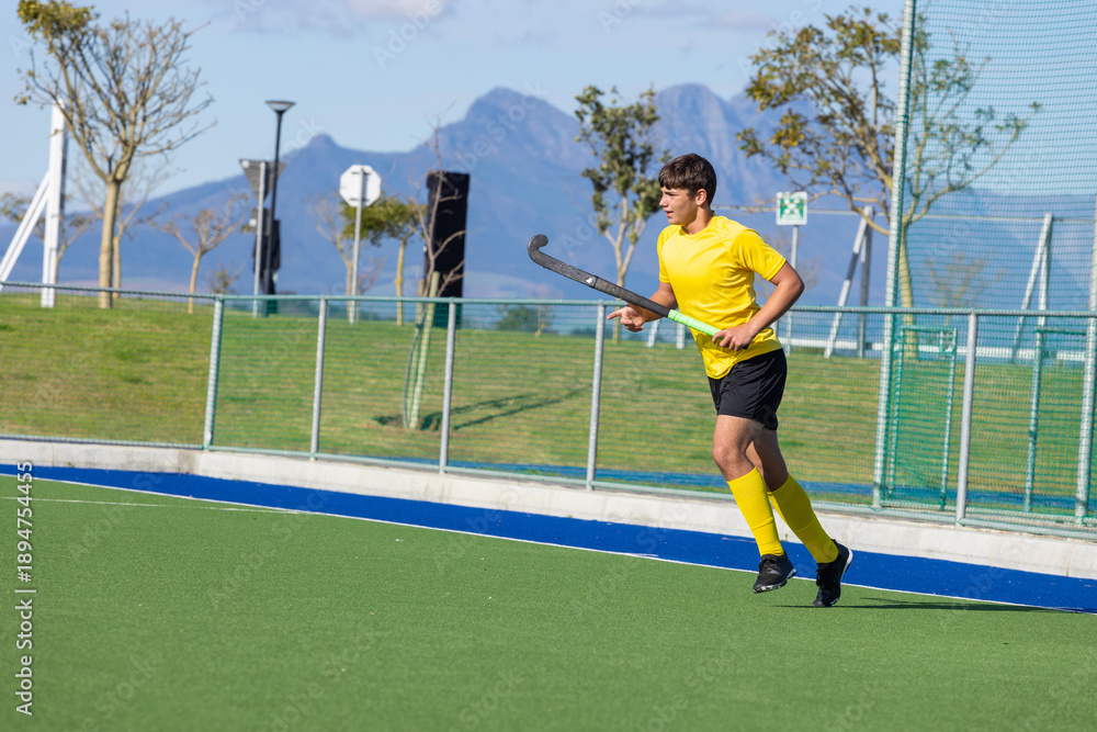 Obraz premium Field hockey player running on bright green turf holding stick next to metal fence and netting
