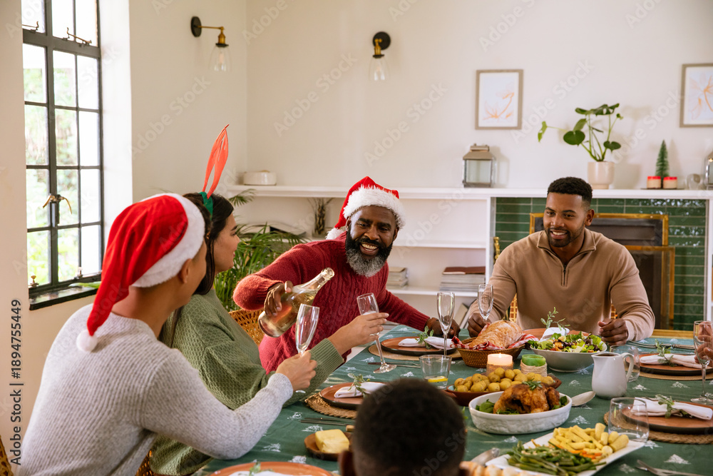 Naklejka premium Diverse family in santa hats at dining table and african american man pouring champagne