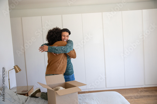Diverse family hugging in bedroom near moving box on gray bedding with brass lamp and smartphone