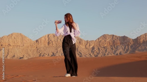 Teen caucasian girl standing on sand dune taking selfie with smartphone in desert during daytime, expressing freedom, travel and social media lifestyle.