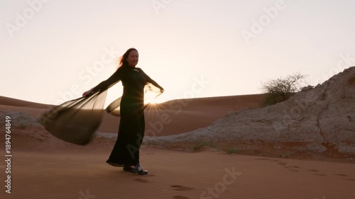 Adult woman in long black dress walking through desert dunes at sunset, backlit by low sun with flowing fabric, expressing freedom, spirituality and inspirational travel mood.