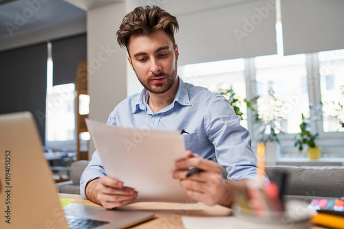 Young entrepreneur carefully reading legal contract and terms of service at desk in modern bright home office.