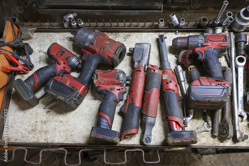 Gritty collection of dirty, red and black cordless power tools on metal workbench. Well used equipment including an impact wrench and drill for automotive repair in garage