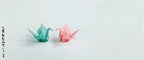 Origami paper cranes made from recycled paper in pastel teal and pink, positioned to face each other against a white background